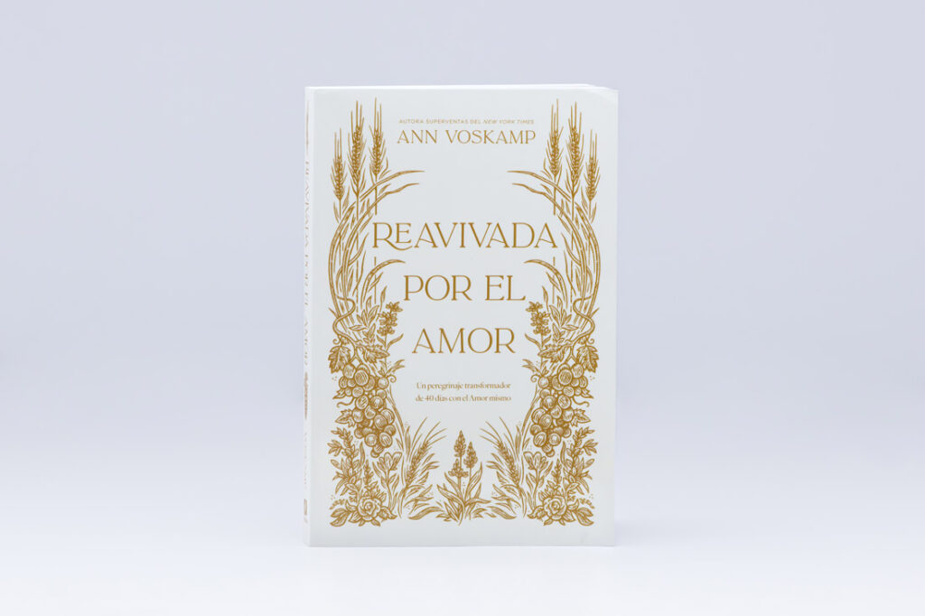 White softcover book with gold foil stamping featuring ornate wheat and floral border design framing the title 'REAVIVADA POR EL AMOR' by Ann Voskamp.