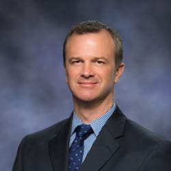 Professional headshot of Marc Paquin, a man with short brown hair wearing a dark suit, blue dress shirt, and blue patterned tie, photographed against a soft blue-gray background.