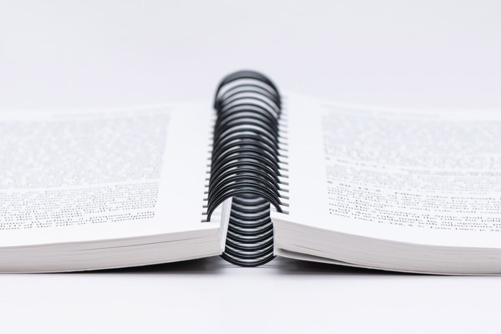 Close-up of spiral bound book or notebook showing black coil binding with white textured covers laying open flat on a white surface.
