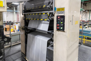 Timsons ZMR web coldset offset printer with large paper web roll feeding through press with yellow and black safety striping and control panel.