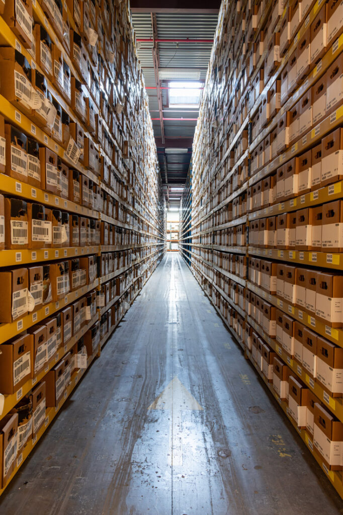 Long narrow aisle between tall shelving units stacked floor to ceiling with labeled cardboard boxes in warehouse with overhead lighting.