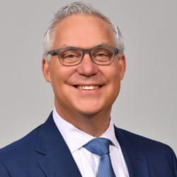 Professional headshot of Todd Mazuka, a man with gray hair wearing black-framed glasses, a navy blue suit, and blue striped tie, smiling against a neutral gray background.