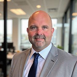 Professional headshot of Steve Zweber, a man wearing a light blue and white checkered button-down shirt, photographed in an office setting with glass walls and bright lighting in the background.