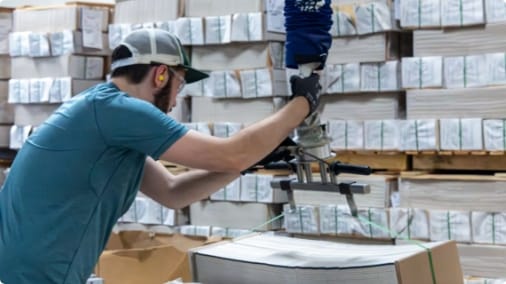 Man in blue shirt operating a robotic arm to handle bundled book pages in a print plant warehouse with stacked materials in background.
