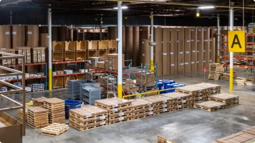 rint manufacturing warehouse interior with rows of large paper rolls, stacked wooden pallets, metal shelving units, and yellow safety signage.