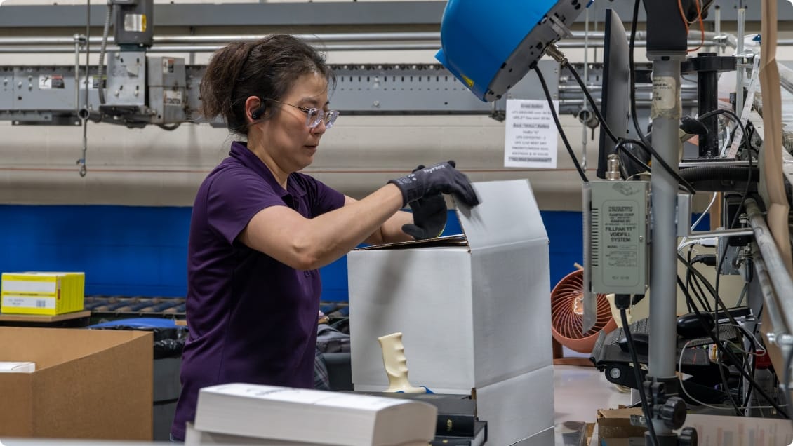 Female worker in purple shirt and safety glasses packing a white box with industrial equipment and cardboard boxes in background.