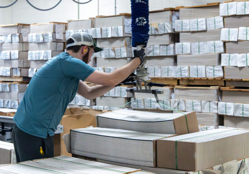 house worker in blue shirt and cap operating an overhead vacuum lift to move stacks of bound books, with pallets of boxed inventory visible in the background.